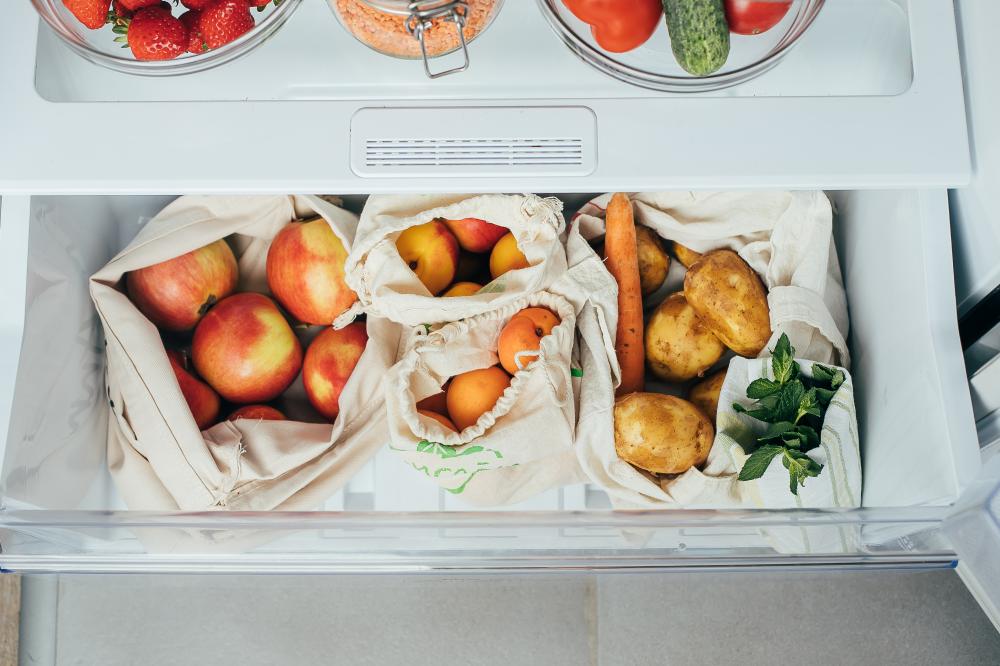 Under counter fridge filled with fresh fruits and vegetables, including apples, apricots, carrots, and potatoes in reusable bags, showcasing organization and functionality for kitchen storage.