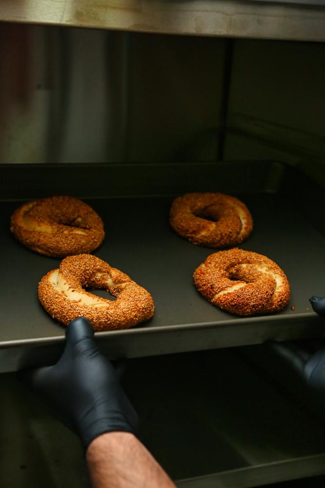Person in black gloves placing freshly baked sesame bagels on an oven tray.