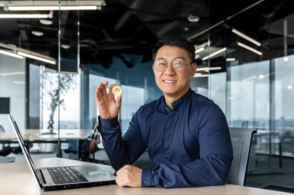 Asian businessman holding a cryptocurrency coin, seated at a laptop in a modern office setting, representing digital marketing and investment strategies for Seattle businesses.