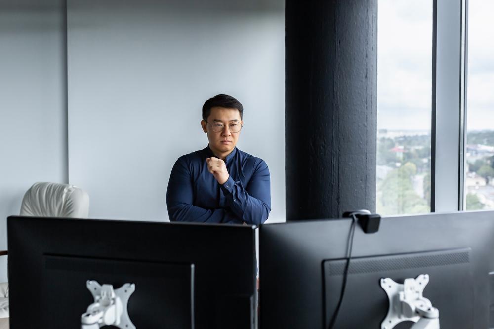 Asian engineer thoughtfully analyzing data on dual monitors in a modern office setting, reflecting the strategic approach of Cloud 7 Agency's SEO services in Seattle.